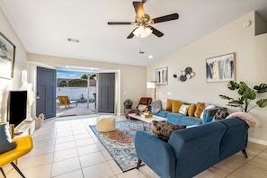A living room with a blue sofa, a coffee table, and a ceiling fan. There is a door leading to an outdoor area with chairs. - Relaxing 4BR Retreat w/ Refreshing Pool (Indio)