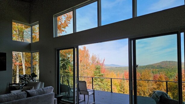 Ein geräumiges Wohnzimmer mit einem großen Schiebeglasfenster, das auf einen Balkon mit herbstlichem Blätterwerk und Hügellandschaft führt.