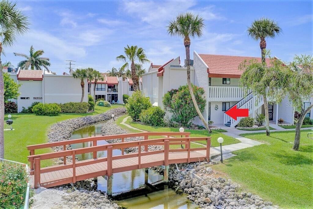 A resort with a wooden bridge over a small water feature, surrounded by palm trees and well-maintained lawns.
