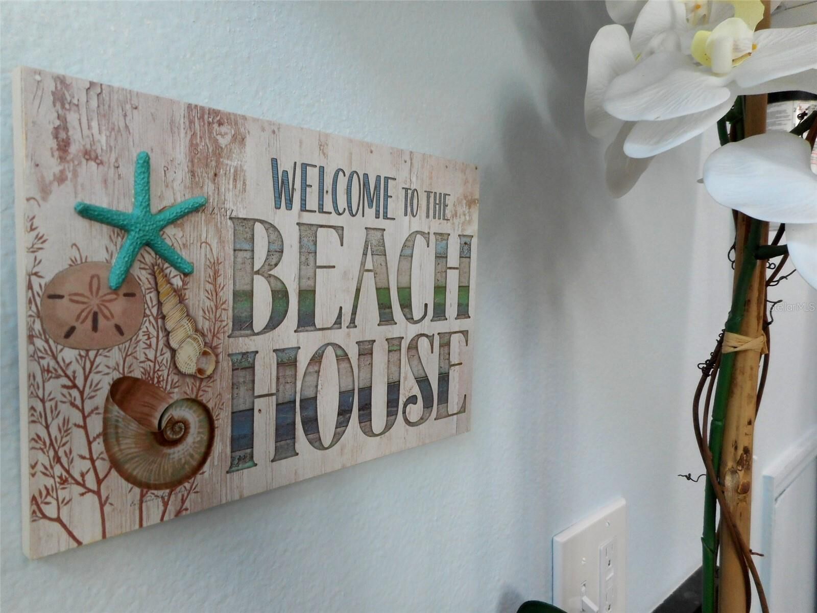 Letrero de bienvenida con el texto 'WELCOME TO THE BEACH HOUSE', decorado con conchas marinas y una estrella de mar.