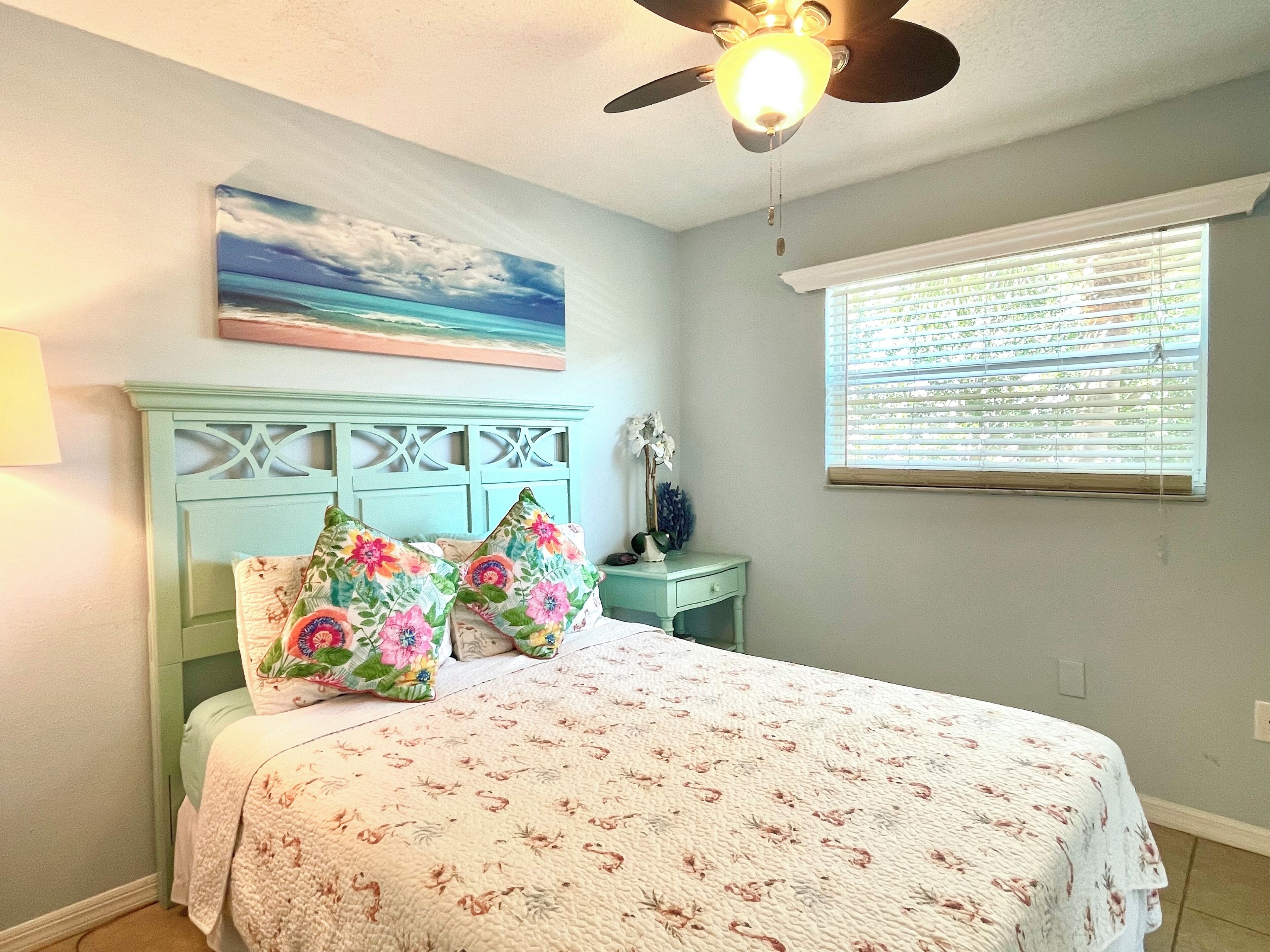 Une chambre à coucher avec un lit, un ventilateur de plafond, une fenêtre avec des stores et un tableau accroché au mur.