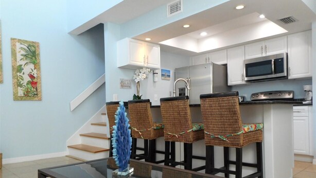 A modern kitchen with white cabinets, stainless steel appliances, and a central island with bar stools. - Seaside Escape (Bradenton Beach)