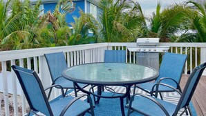 A round glass table with four blue chairs, a grill, and a view of a blue building with palm trees. - Seaside Escape (Bradenton Beach)