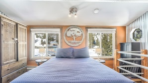 Un dormitorio con una cama grande, armario de madera, dos ventanas que dan a un paisaje nevado y un adorno de árbol colgado en la pared.