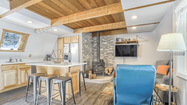 Una cocina moderna con vigas de madera, una isla central, taburetes de bar, una pared de piedra, un televisor y una silla azul.