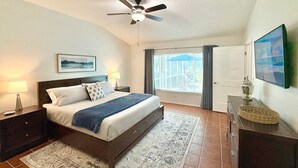 A bedroom with a bed, nightstands, a ceiling fan, a window with curtains, a door, and a wall-mounted TV. - The Salty Pelican (Bradenton)
