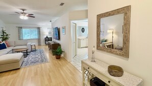 A spacious living room with a ceiling fan, a sofa, a coffee table, and a TV. There is a hallway leading to a kitchen and a dining area. - The Salty Pelican (Bradenton)