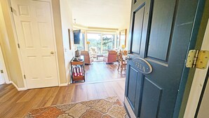 Um corredor com piso de madeira, uma porta azul escura com placa de número e vista para uma sala de estar com sofá e mesa de centro.