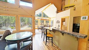 A spacious dining area with a large window overlooking a forest, a kitchen with wooden cabinets, and a bar area with a digital thermometer. - Aspen Place River Access & Ping Pong (Bend)