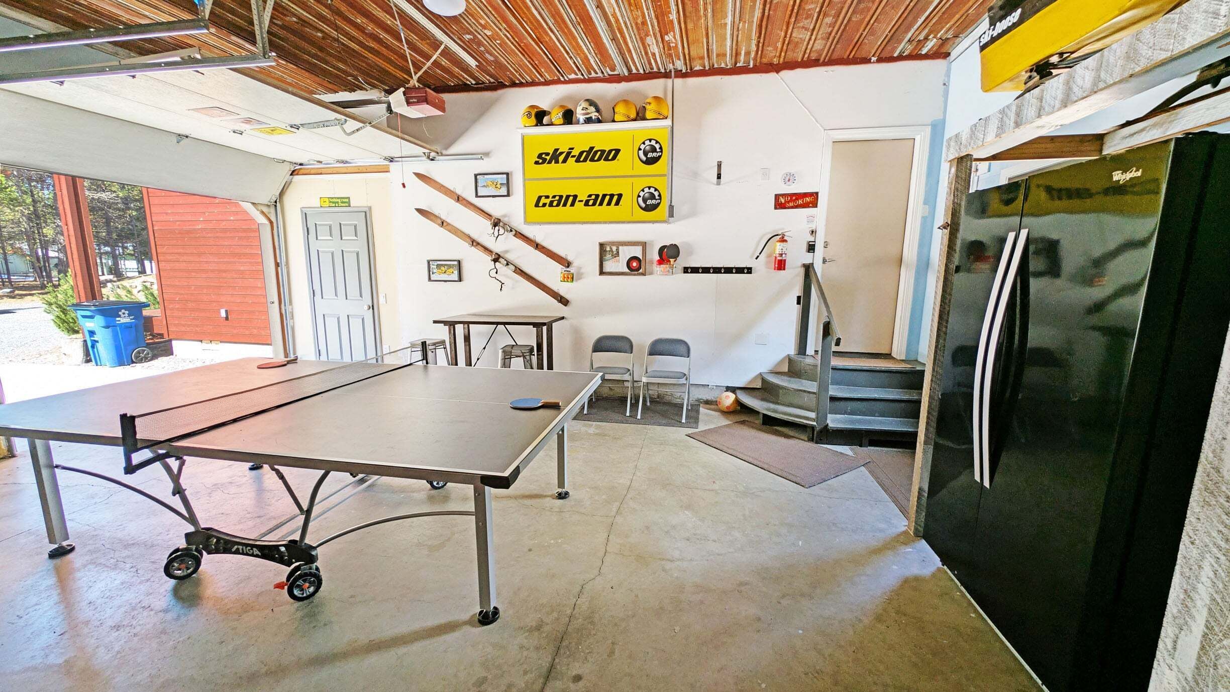 Une pièce équipée d’une table de ping-pong, de chaises, d’un réfrigérateur et d’une enseigne Can-Am représentant un véhicule de motoneige.