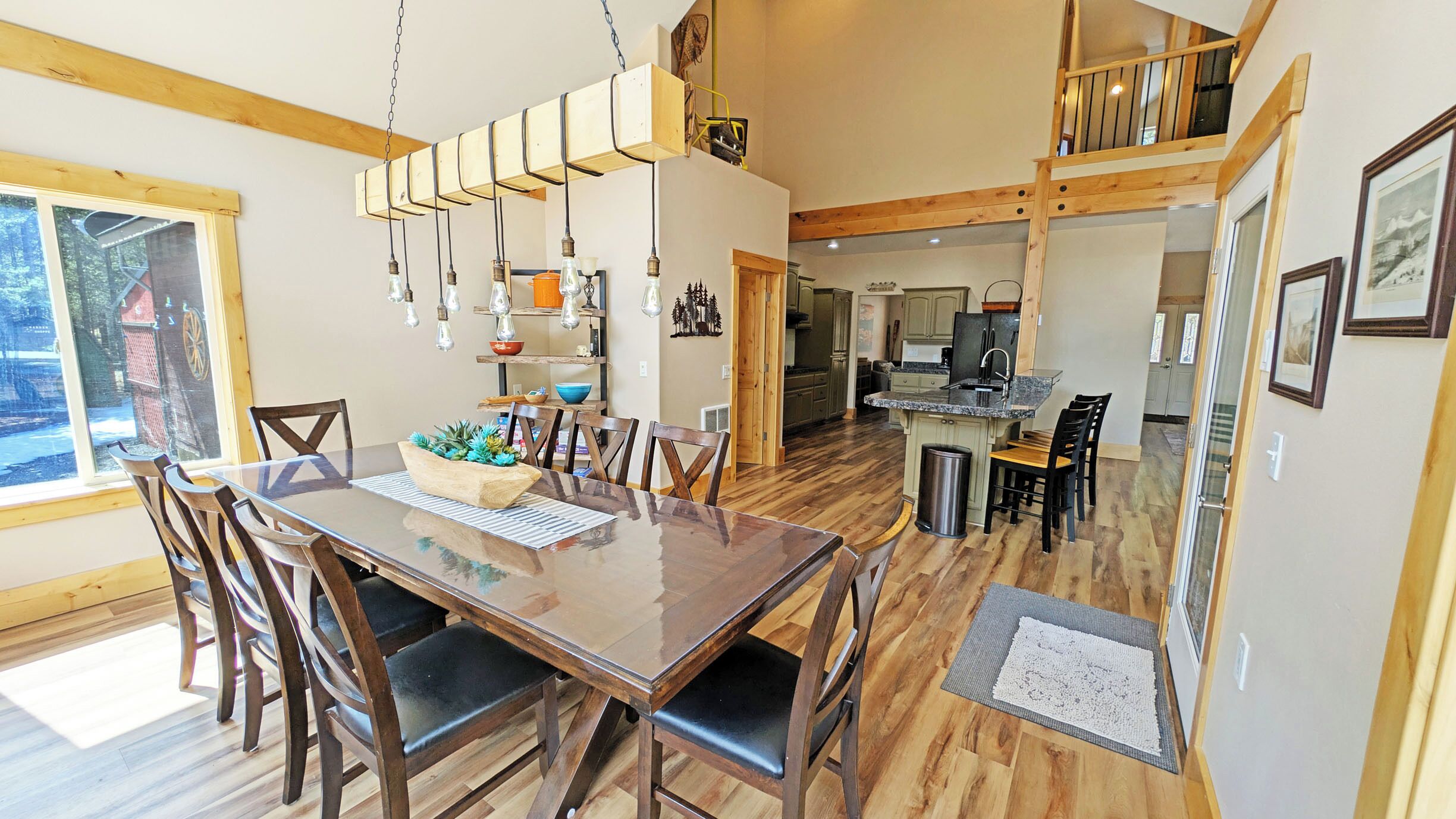 Uma sala de jantar espaçosa com mesa e cadeiras de madeira, uma cozinha ao fundo e vista para o exterior por uma janela grande.