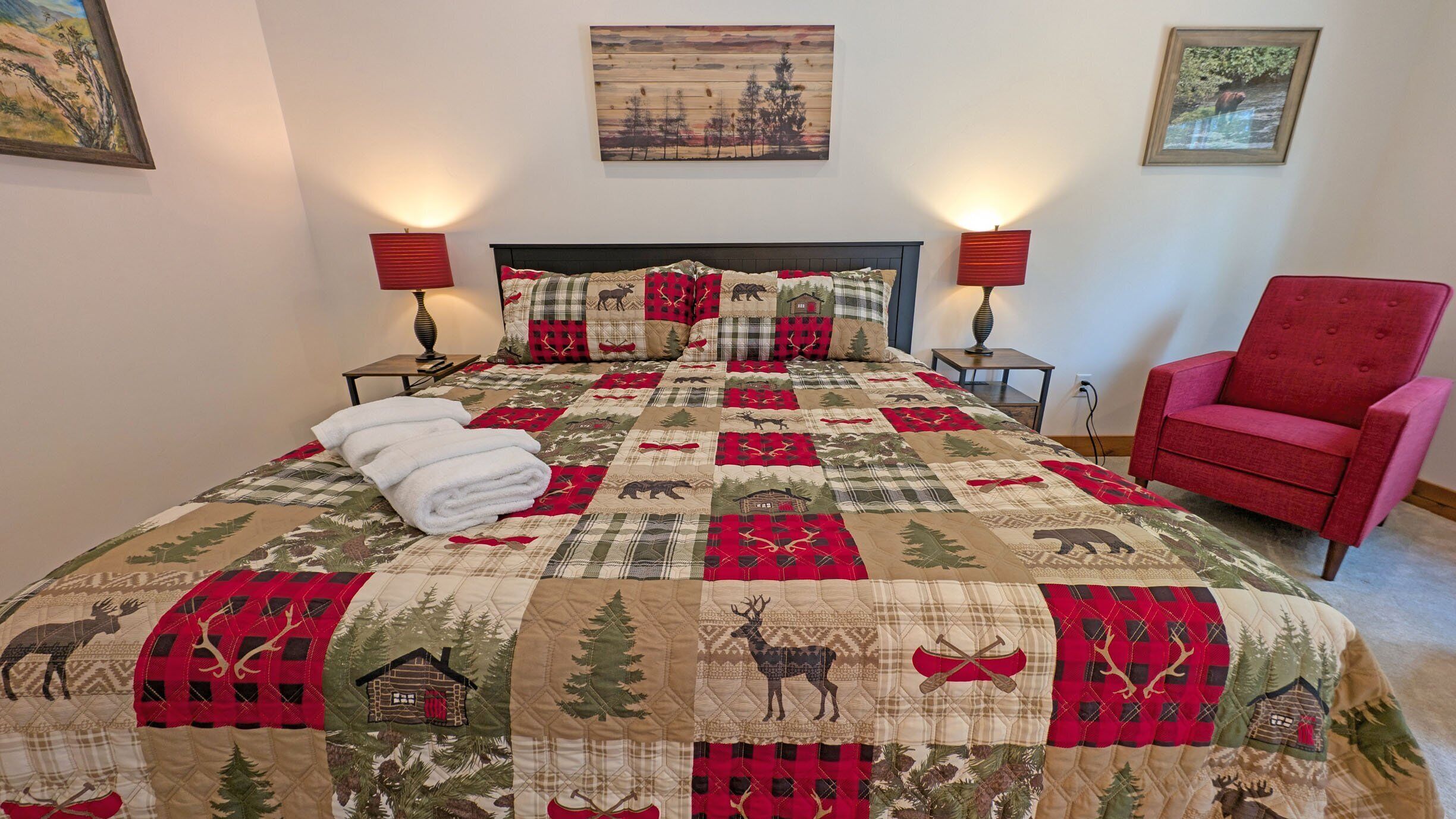 A bed with a quilt featuring forest and wildlife patterns, two bedside lamps, a red chair, and a painting on the wall.