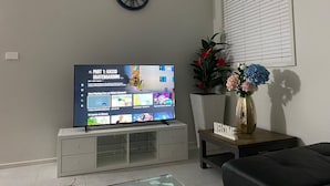 A living room with a flat-screen TV displaying a sports channel, a decorative clock, a potted plant, and a welcome sign on a side table. - Spacious Family & Group Retreat | Sleeps 10 | 3 Baths | free parking  (North Kellyville)