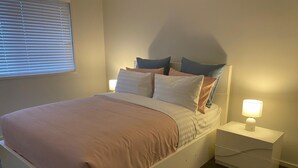 A bedroom with a bed, pillows, a bedside table with a lamp, and a window with blinds. - Spacious Family & Group Retreat | Sleeps 10 | 3 Baths | free parking  (North Kellyville)