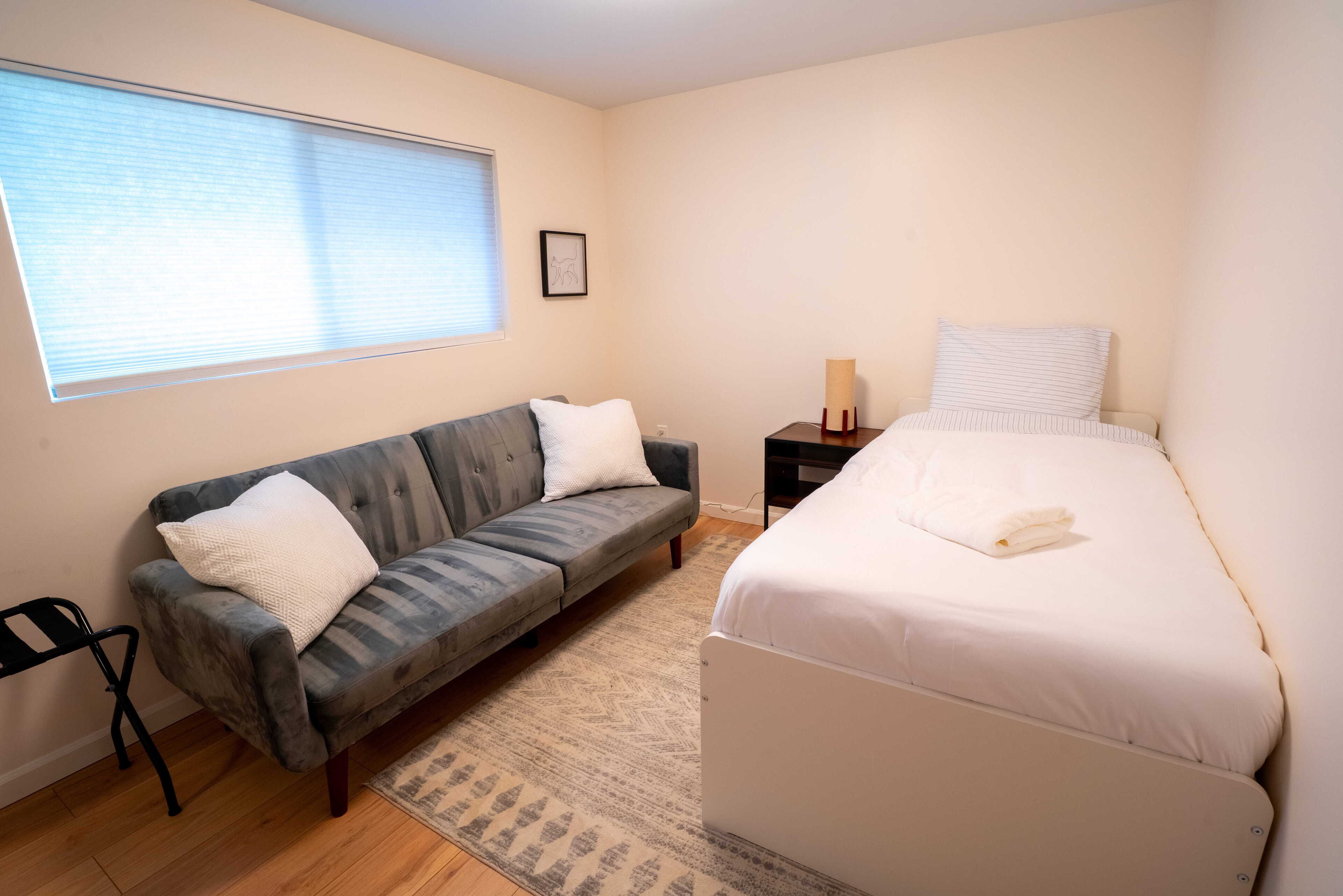 A bedroom with a grey sofa, a white bed, a small bedside table with a lamp, and a window with blinds.