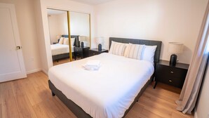Quarto bem arrumado com uma cama grande, uma mesinha de cabeceira com abajur, um espelho e um guarda-roupa.