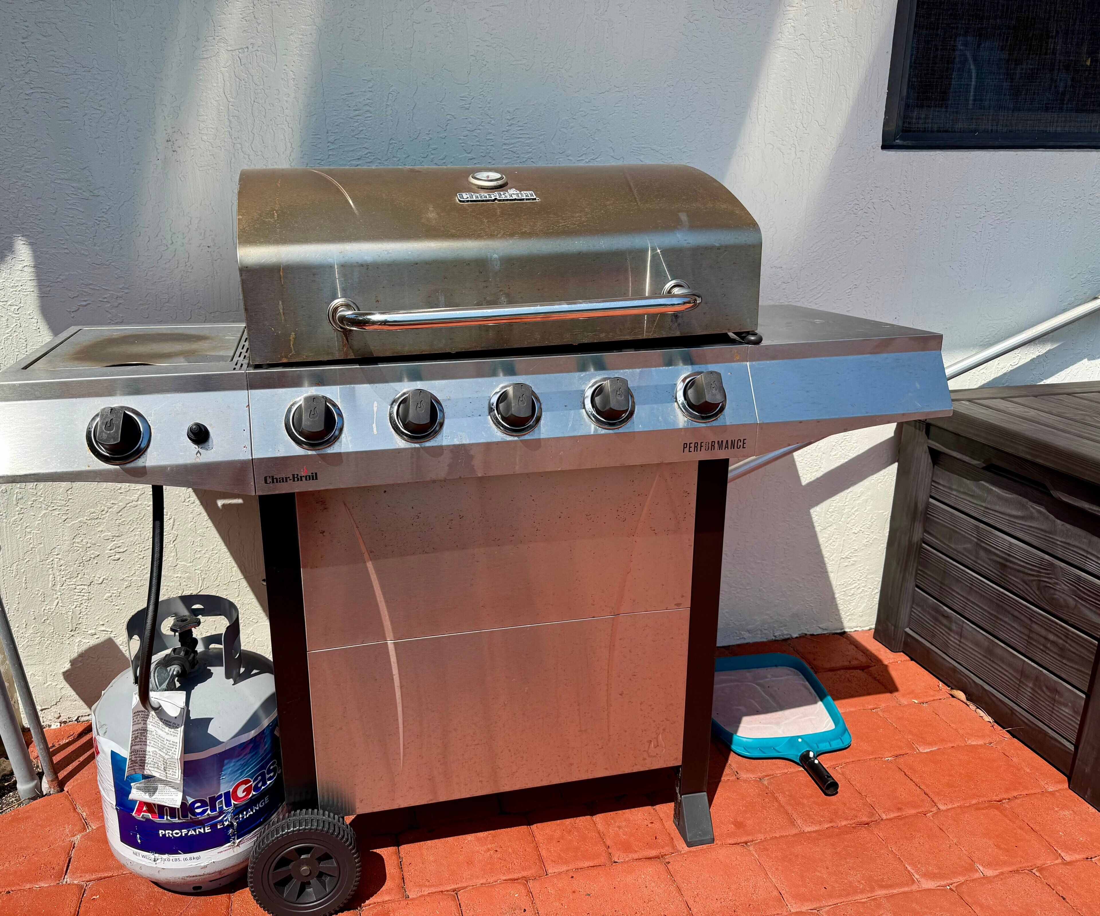 Una parrilla de acero inoxidable con tanque de gas y una parrilla de carbón debajo, ubicada sobre un patio de ladrillo.
