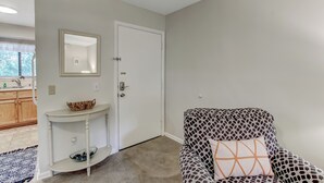 A room with a white door, a small white console table with a basket, a patterned armchair with a cushion, and a blue patterned rug. - 10D Amelia Landings (Fernandina Beach)