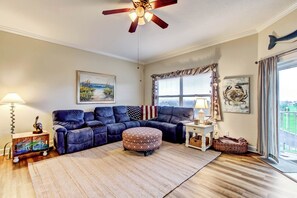 A living room with a sectional sofa, ottoman, ceiling fan, artwork, and a view through a window. - 1 Block From the Beach, Next to Ft Clinch State Park and 2 Miles From Historic Centre Street (Fernandina Beach)