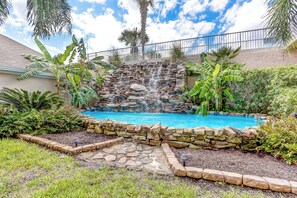 A backyard with a stone waterfall, a swimming pool, and lush greenery including banana trees and ferns. - 1 Block From the Beach, Next to Ft Clinch State Park and 2 Miles From Historic Centre Street (Fernandina Beach)