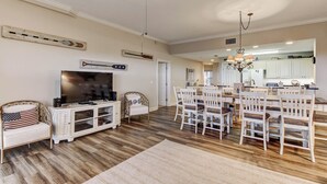 A spacious living room with a large TV, a dining area with white chairs, and a kitchen in the background. - 1 Block From the Beach, Next to Ft Clinch State Park and 2 Miles From Historic Centre Street (Fernandina Beach)