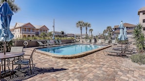 A pool area with lounge chairs and umbrellas, surrounded by palm trees and a building with a balcony. - 109 Oceans of Amelia (Fernandina Beach)