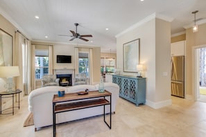 A living room with a fireplace, a ceiling fan, a TV, and a kitchen area with a refrigerator. - 1016 San Fernando Street (Fernandina Beach)