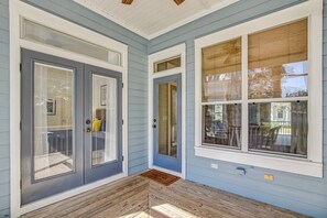 Een overdekt terras met een houten vloer, een blauwe deur met een ruit en een raam met een houten zonwering.