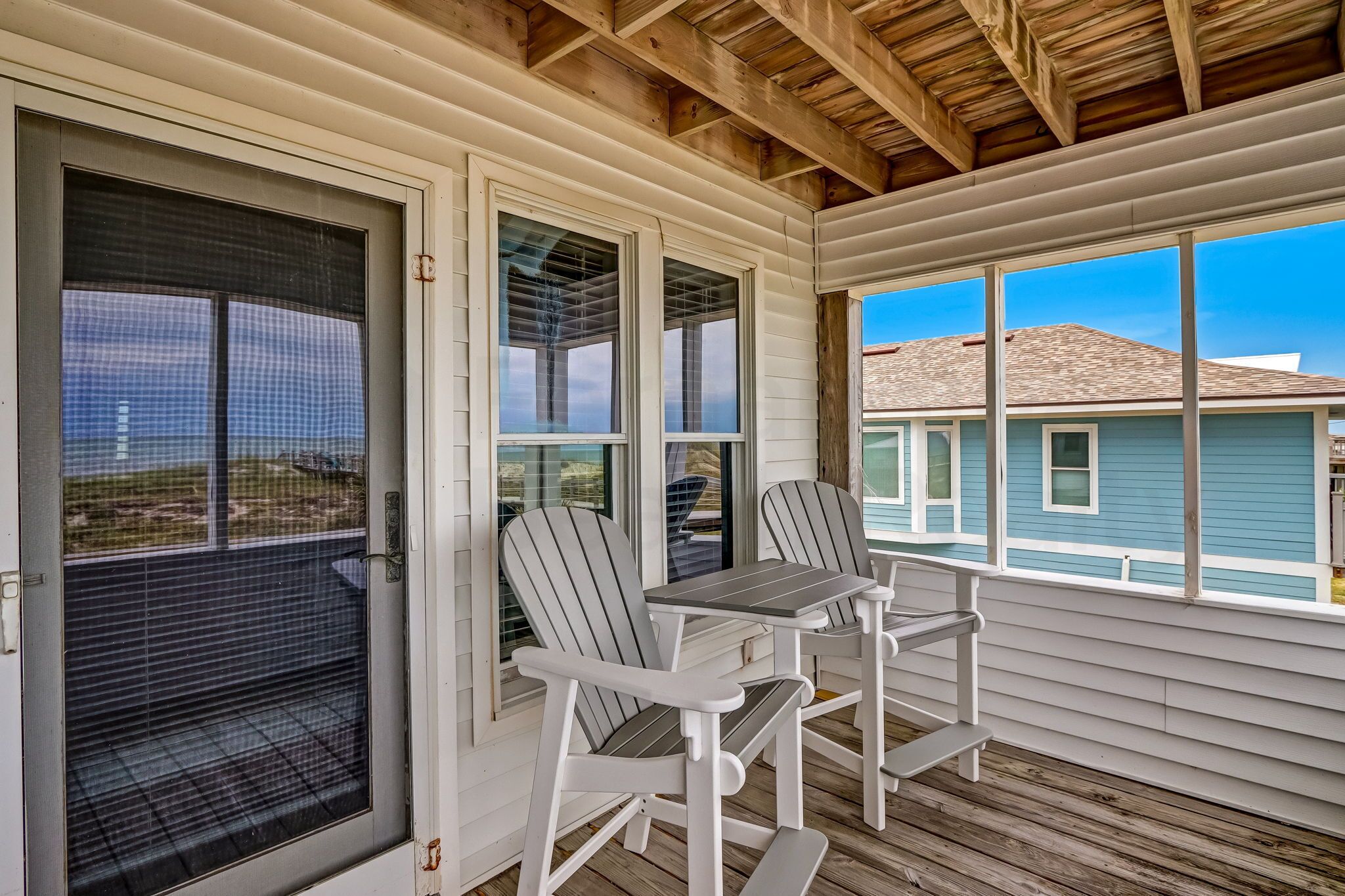 A covered porch with two white Adirondack chairs and a table, overlooking a blue house with a tiled roof.