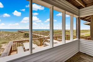 Ein überdachter Außenbereich mit Holzdeck, weißen Geländern und Blick auf Strand und Dünen.