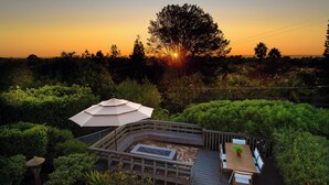 Une terrasse avec une table et des chaises, entourée d’une végétation luxuriante, avec un coucher de soleil en arrière-plan.