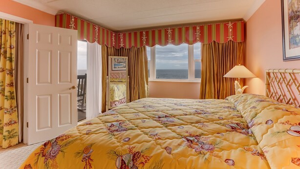 A bedroom with a bed, a window with curtains, a door, and a lamp. - 446 Amelia By The Sea (Fernandina Beach)