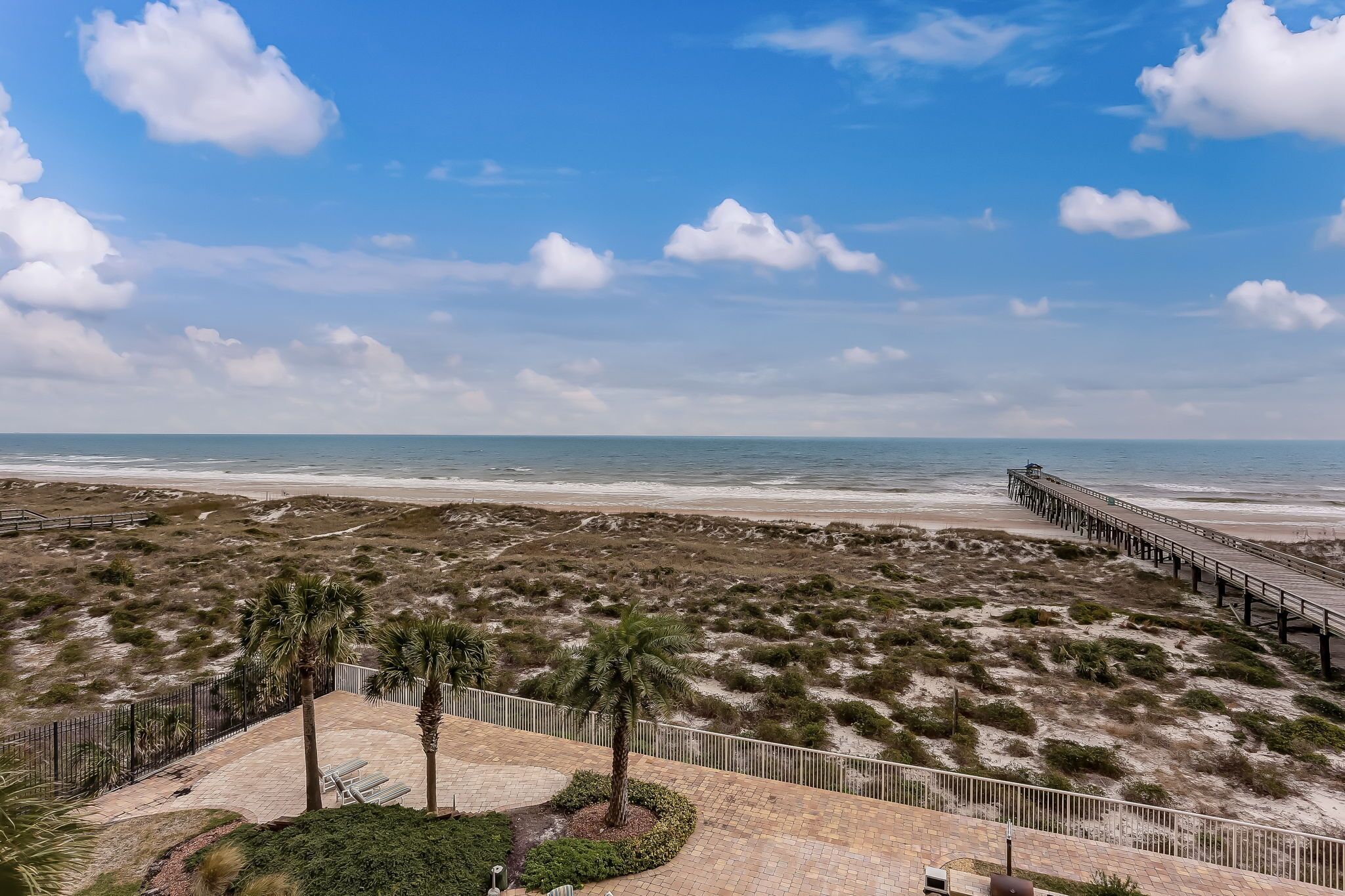 Une plage avec vue sur une jetée en bois qui s’avance dans l’océan, des dunes de sable et une zone délimitée par une clôture, où se trouvent des palmiers.