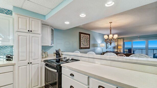 A kitchen with white cabinets, a black stove, and a large island with drawers. There is a chandelier and a window with a view. - 438 Amelia By The Sea (Fernandina Beach)