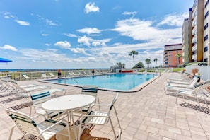 Un espace aménagé au bord de la piscine, avec des chaises longues et une table, un ciel d’un bleu éclatant et un bâtiment en arrière-plan.