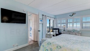 A bedroom with a bed, a TV mounted on the wall, a window with blinds, and a door leading to another room. - 438 Amelia By The Sea (Fernandina Beach)