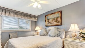 A bedroom with a large bed, two bedside tables with lamps, a ceiling fan, a window with blinds, a painting on the wall, and a decorative vase on the nightstand. - 210 Oceans of Amelia (Fernandina Beach)