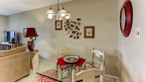 A dining area with a glass table, red napkins, and a bowl. There are chairs, a lamp, and framed pictures on the wall. - 208 Sand Dollar (Fernandina Beach)