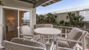 A covered outdoor seating area with a round table and white chairs, overlooking a building with palm trees and a clear sky. - 208 Sand Dollar (Fernandina Beach)