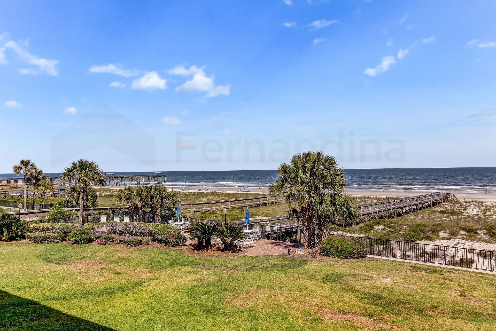 A beach view with a wooden boardwalk, palm trees, and a grassy area.