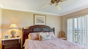 A bedroom with a large bed, a nightstand with a lamp, a wooden chair, and a ceiling fan. - 204 Atlantis (Fernandina Beach)