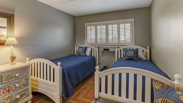 Une chambre à coucher avec deux lits en bois blancs, chacun agrémenté de draps bleus et de coussins décoratifs. On y trouve une commode dont le devant du tiroir est orné d’un motif de poisson, une fenêtre avec des stores et une horloge au mur.