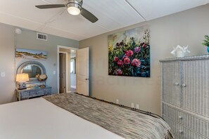 A bedroom with a bed, a ceiling fan, a painting, a wicker cabinet, and a door leading to another room. - 203 Oceans of Amelia (Fernandina Beach)