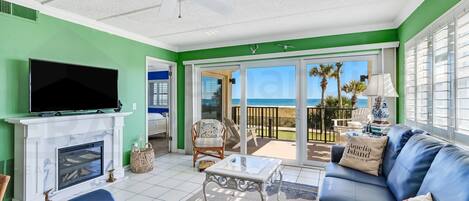 A living room with a fireplace, a television, a sofa, and a balcony with a view of the ocean.
