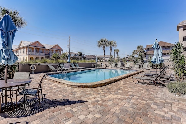 A swimming pool with lounge chairs and umbrellas, surrounded by palm trees and a building with a balcony.