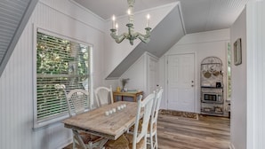 A dining area with a wooden table and white chairs, a chandelier, and a view through a window with blinds. - 313 New Street (Fernandina Beach)