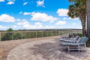A sunlit outdoor lounge area with a curved brick patio, a palm tree, and a view of a bridge in the background. - 334 Amelia By The Sea (Fernandina Beach)
