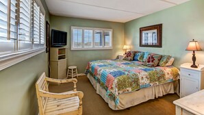 Ein Schlafzimmer mit einem Bett, einem Sessel, einem Schreibtisch, einer Lampe, einem Fernseher und einem Fenster mit Jalousien.