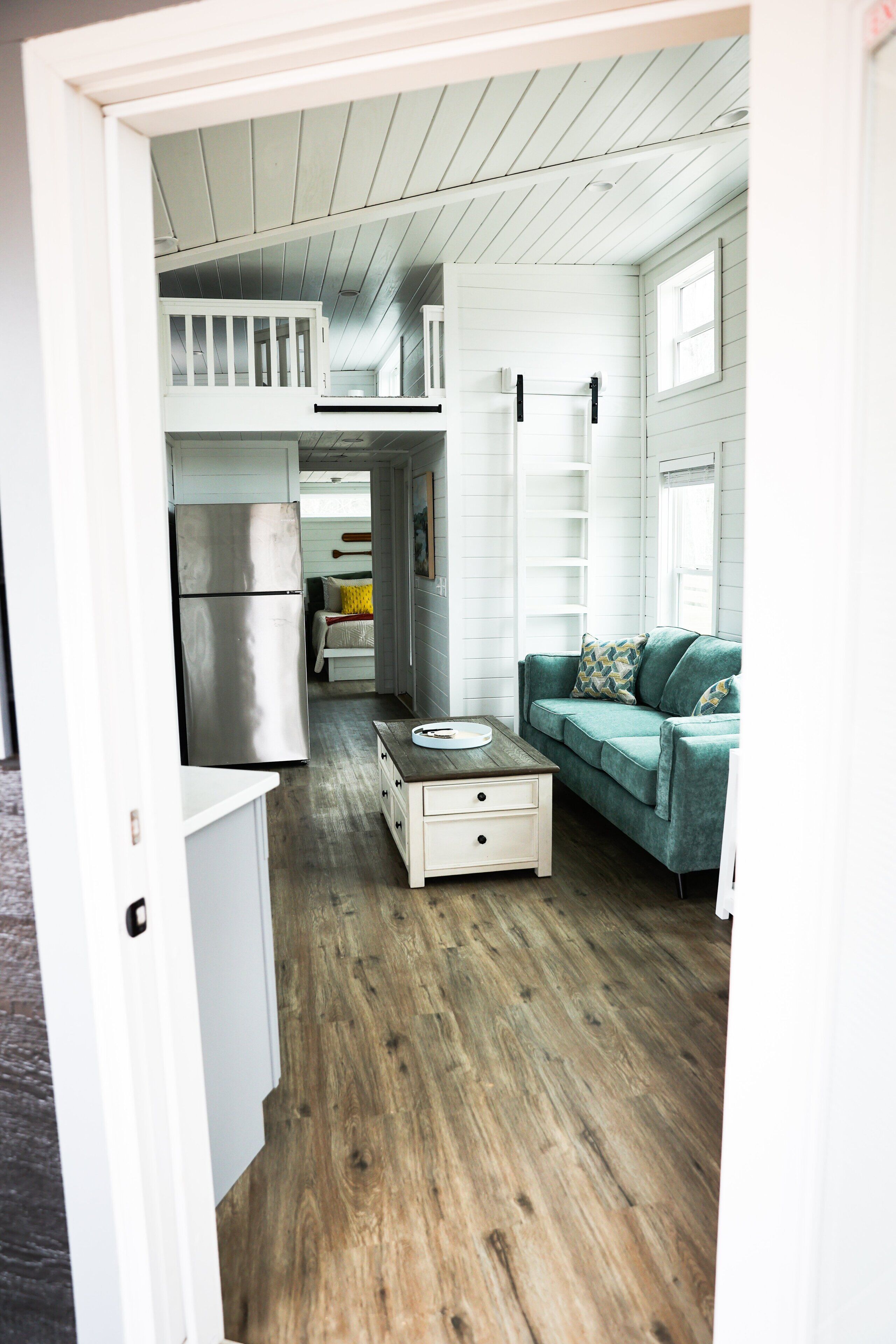 A narrow hallway with a wooden floor, a green sofa with patterned cushions, a white dresser, and a stainless steel refrigerator.