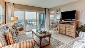 A spacious living room with a large flat-screen TV, striped sofas, a glass coffee table, and a view of the balcony. - 7th Floor, Corner Property on the top Floor With Easy Beach Access Exclusive Fishing Pier (Fernandina Beach)